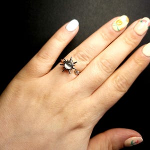 May include: A silver ring with a sunburst design and a central clear, round stone. The ring is on a hand with white nail polish and floral nail art. The background is black.