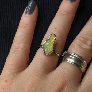 Op de afbeelding: Een zilveren ring met een peervormige, lichtgroene edelsteen. De edelsteen is gezet in een zilveren draadframe. Een andere ring met een gouden band is ook zichtbaar. De achtergrond is donker.