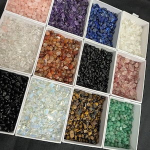 May include: A selection of small, polished gemstones in white rectangular containers. The stones are various colours, including pink, purple, blue, white, orange, black, and green. The arrangement is on a black surface.