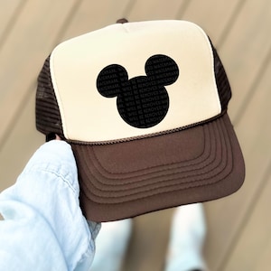 May include: A brown and tan trucker hat featuring a black Mickey Mouse silhouette on the front. The hat has a mesh back and a curved brim. The text "WATERMARK WILL BE REMOVED WATERMARK" is printed on the Mickey Mouse silhouette.