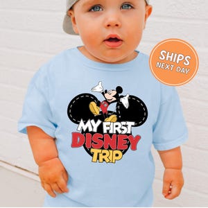 A baby wearing a light blue t-shirt with a Mickey Mouse graphic and text that says "My first Disney trip".