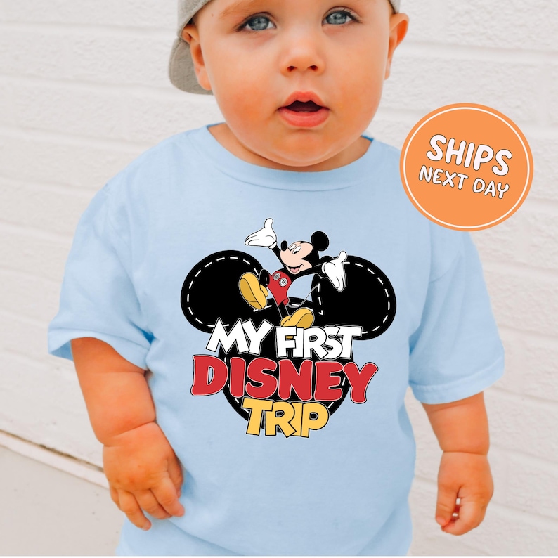 A baby wearing a light blue t-shirt with a Mickey Mouse graphic and text that says "My first Disney trip".