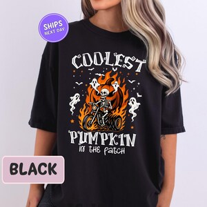 May include: Black t-shirt with a Halloween-themed graphic. The design features a skeleton riding a motorcycle inside a flaming pumpkin, with the text "COOLEST PUMPKIN in the patch". White ghosts and stars surround the central image.