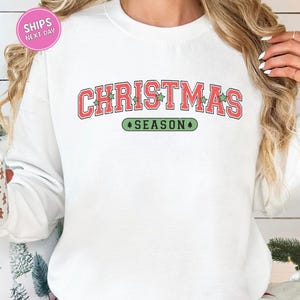 May include: White sweatshirt with the words "CHRISTMAS SEASON" in red and green lettering. The word "CHRISTMAS" is arched across the top, and "SEASON" is below. The sweatshirt has a crew neck.