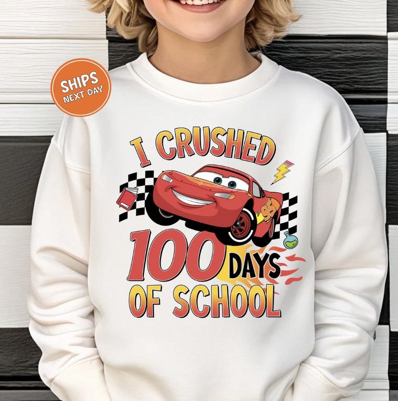 A person wearing a white sweatshirt with a graphic design featuring a red car and the text "I CRUSHED 100 DAYS OF SCHOOL".