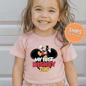 A young girl wearing a pink t-shirt with a Mickey Mouse graphic and text, smiling at the camera.