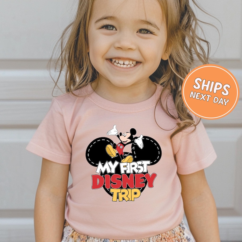 A young girl wearing a pink t-shirt with a Mickey Mouse graphic and text, smiling at the camera.