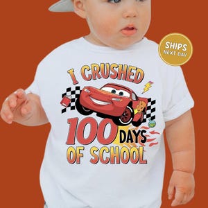A baby wearing a white t-shirt with a graphic of a red car and the text "I CRUSHED 100 DAYS OF SCHOOL" printed on it.