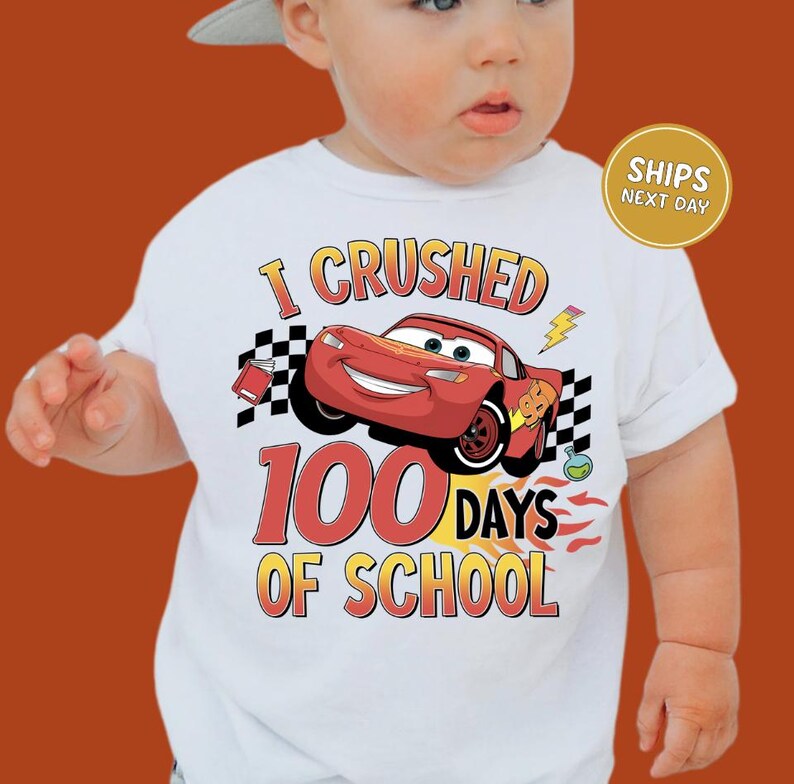 A baby wearing a white t-shirt with a graphic of a red car and the text "I CRUSHED 100 DAYS OF SCHOOL" printed on it.