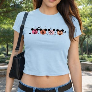 May include: A light blue short-sleeved t-shirt featuring a graphic print of four pumpkins with mouse-ear silhouettes in shades of pink and orange, accented by small black bats and star shapes arranged in a horizontal line across the chest.