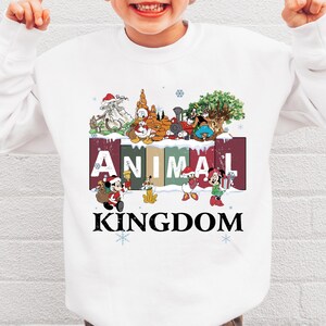 A white sweatshirt with a colorful graphic design featuring various Disney characters and the word "ANIMAL KINGDOM" printed on it.