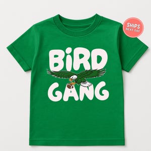 May include: Green t-shirt with the words "BIRD GANG" in white block letters. A cartoon eagle with green and white feathers and a football is centered between the words. The shirt is short-sleeved.