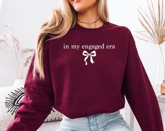May include: A burgundy sweatshirt with the text "in my engaged era" in white, along with a white bow graphic. The long-sleeved top is made of a soft material and is suitable for casual wear.