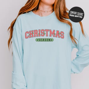 May include: A light blue long-sleeve shirt with the words "CHRISTMAS SEASON" in red and green lettering. The shirt has a crew neck and is made of a soft material. The text is in a collegiate style font.