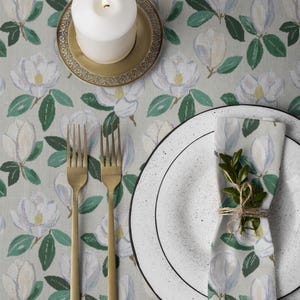 May include: A table setting featuring a white candle on a gold-colored plate, two gold forks, and a white plate with a black rim. A floral napkin with a sprig of greenery is tied with twine. The tablecloth has a magnolia flower pattern.