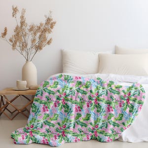 May include: A blanket with a floral pattern of pink holly berries, green leaves, and light blue accents. The blanket is draped over a bed with white linens. The design creates a cosy and inviting aesthetic.