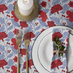 May include: A white tablecloth with a red and blue floral pattern, a white pillar candle on a gold plate, two gold forks, and a white speckled plate with a matching floral napkin tied with twine and a small green sprig of leaves.