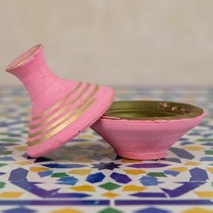 Puede incluir: Un cuenco decorativo rosa con tapa a juego. La tapa está adornada con rayas doradas. El interior del cuenco es dorado. El cuenco y la tapa descansan sobre una superficie de baldosas coloridas.