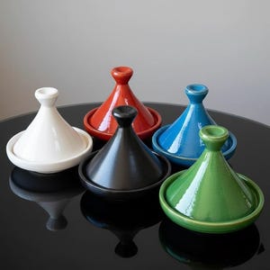 Peut inclure: Plusieurs tagines en céramique colorées. Les tagines sont de différentes couleurs : blanc, rouge, noir, bleu et vert. Chaque pot a un couvercle conique et une base peu profonde. Les pots sont disposés sur une surface noire réfléchissante.