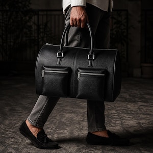 May include: A black leather duffel bag with two front zippered pockets and silver-tone hardware. The bag has two top handles and a cylindrical shape. The bag is being carried by a person wearing black loafers.