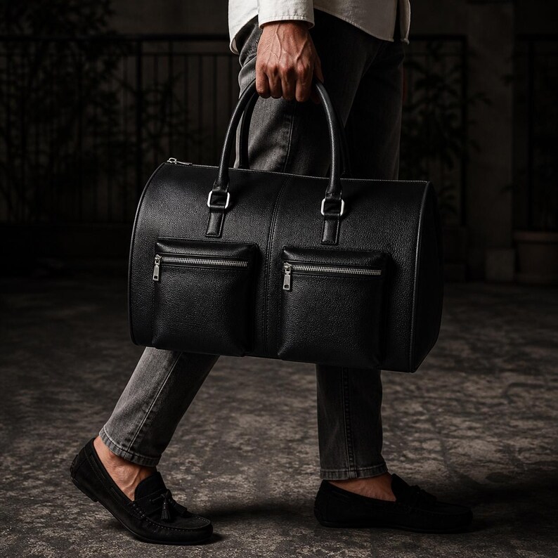 May include: A black leather duffel bag with two front zippered pockets and silver-tone hardware. The bag has two top handles and a cylindrical shape. The bag is being carried by a person wearing black loafers.
