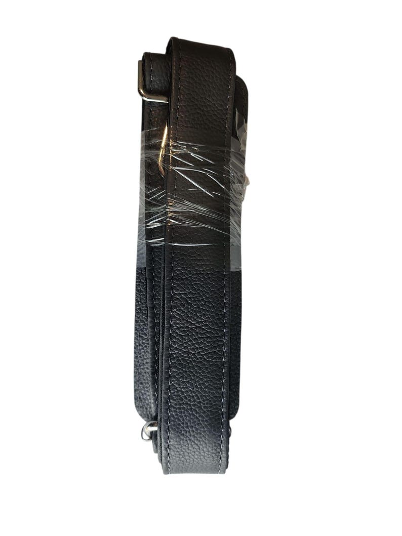 May include: A black, textured leather bag strap with silver-colored metal hardware. The strap is wrapped in clear plastic. The strap is designed to be adjustable and is likely for a shoulder bag or purse.