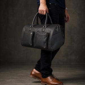 May include: A black leather duffel bag with two front zippered pockets and a wraparound zipper. The bag has two top handles and silver-colored hardware. The person is wearing black pants and brown leather shoes.
