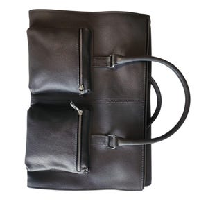 May include: Dark brown leather briefcase with two zippered pockets on the front. The briefcase has a handle and a buckle closure. The bag is made of a textured leather material.
