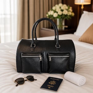 May include: A black leather duffel bag with two front zippered pockets, sitting on a white bedspread. Sunglasses, a rolled white towel, and a U.S. passport are next to the bag. The bag has two top handles.