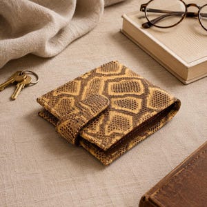 May include: A brown and tan snakeskin-patterned wallet with a snap closure. The wallet is open, revealing a glimpse of the interior. A set of keys and a pair of spectacles rest nearby on a neutral-toned surface.
