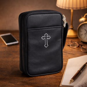 May include: Black rectangular Bible cover with a white embroidered cross. The cover has a zipper closure and a small handle. The cover is made of a smooth material and has a pocket on the front. The background includes a lamp, clock, and notebook.