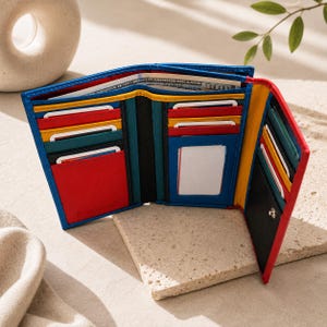 May include: A multi-colored leather trifold wallet featuring blue, red, yellow, and black panels. The interior includes multiple card slots, a clear ID window, a bill compartment containing a 100 dollar bill, and the embossed brand name Pompah.