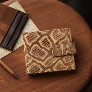 Può includere: Un portafoglio quadrato con motivo a pelle di serpente beige e marrone, con chiusura a scatto. Il portafoglio è su una superficie di legno con una penna e un porta carte in pelle marrone sullo sfondo. È un piccolo accessorio compatto.