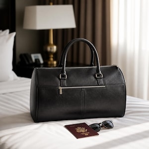 May include: A black leather duffel bag with silver zippers and handles sits on a white bed. A passport and sunglasses are next to the bag. The scene is set in a hotel room with a lamp and curtains in the background.