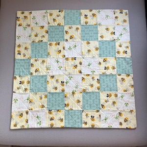 May include: A patchwork quilt featuring a repeating pattern of yellow and black bees, green frogs, and floral designs on a white and yellow background. The quilt also has squares of light blue fabric. The quilt is square in shape.