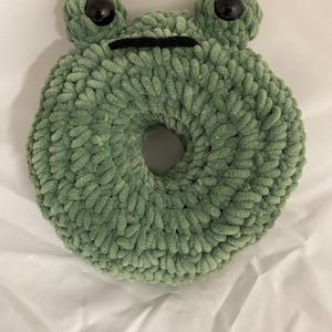 May include: Green crocheted frog-shaped donut pillow with black eyes and a black mouth.