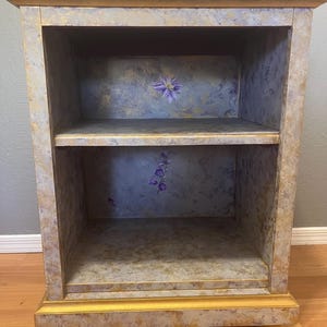 May include: A small, two-shelf bookcase with a gold-painted finish and a distressed, textured look. The shelves are lined with a floral pattern in shades of purple and blue.