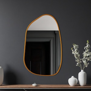 Peut inclure: Un miroir mural de forme irrégulière avec un cadre doré. Le miroir est accroché sur un mur gris foncé au-dessus d'une console en bois. Deux vases blancs et un petit objet décoratif sont sur la table, avec un vase de fleurs blanches à droite.