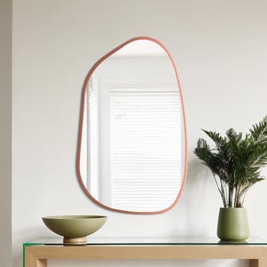 Puede incluir: Un espejo de forma irregular con un marco color salmón cuelga de una pared blanca. Debajo, una mesa con tapa de cristal y marco de madera sostiene un cuenco verde y un jarrón verde con una planta. El ambiente es minimalista y moderno.