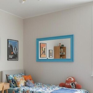 May include: A bedroom with a bed, desk, and artwork. The bed has a floral patterned comforter, pillows, and a blue blanket. A blue-framed mirror hangs above the bed. A desk with a lamp and chair is on the left. A gold light fixture hangs from the ceiling.