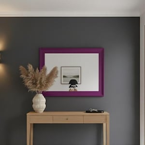May include: An interior shot of a modern entryway with a wooden console table, a decorative vase with dried plants, and a framed mirror with a purple border. A potted plant and a wooden door are also visible.