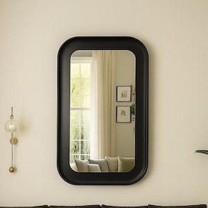 May include: A large, rectangular black framed mirror with rounded corners. The mirror reflects a room with a window, curtains, and a sofa. A gold wall sconce with a glass globe is to the left.