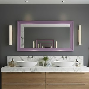 May include: A modern bathroom vanity with a light wood finish, featuring two white vessel sinks and a marble countertop. A large rectangular mirror with a lavender frame hangs above the vanity. Two sconces flank the mirror, and two gray robes hang on the walls.