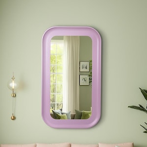 May include: A rectangular, rounded-corner mirror with a light purple frame. The mirror reflects a room with a window, two framed pictures, and a sofa with pillows. The wall is painted a light green color.