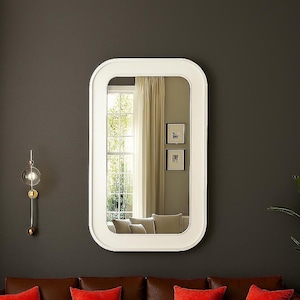 May include: A rectangular white-framed mirror with rounded corners hangs on a dark gray wall. Below, a brown leather sofa with red throw pillows is visible. A decorative wall sconce and a potted plant complete the scene.