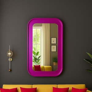 May include: A vibrant magenta-framed mirror hangs on a dark gray wall above a yellow sofa with red pillows. A gold wall sconce and a potted plant add to the room's decor. The mirror reflects a window and additional seating.