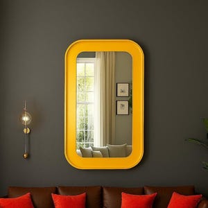 May include: A rectangular yellow mirror with rounded corners hangs on a dark gray wall. The mirror reflects a window with white curtains and a glimpse of a living room. A brown leather sofa with red pillows sits below the mirror.