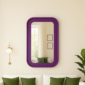 May include: A rectangular purple mirror with rounded corners hangs above a white sofa with green pillows. A gold wall sconce and a potted plant are also visible. The mirror reflects a window and two framed pictures.