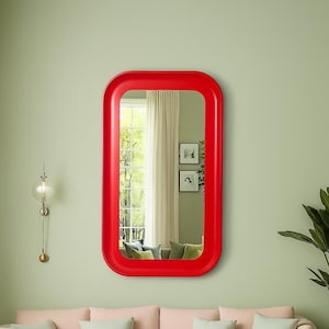 May include: A rectangular red mirror with rounded corners hangs on a pale green wall. The mirror reflects a window with white curtains, two framed pictures, and a sofa with green and beige cushions. A decorative light fixture is to the left.