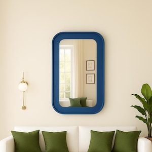 May include: A modern interior scene featuring a navy blue rectangular mirror with rounded corners. The mirror reflects a window, two framed pictures, and green cushions on a white sofa. A gold wall sconce and a potted plant complete the decor.